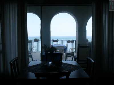 Dalla sala da pranzo intravediamo una veranda delineata da archi in cemento bianco, attraverso cui scorgiamo quel mare che sembra ricreare il paesaggio di uno dei tanti ritratti appesi al muro